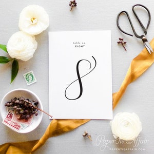 Peut inclure: Carte blanche avec calligraphie noire pour le num&eacute;ro de table huit. La carte est entour&eacute;e de fleurs blanches, d'un ruban dor&eacute; et d'autres &eacute;l&eacute;ments d&eacute;coratifs.