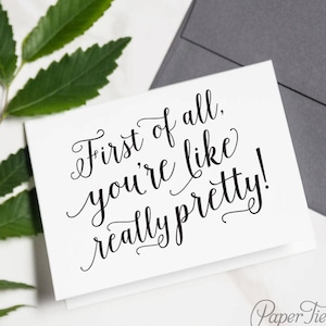 Może przedstawiać: Biała kartka z czarnym kaligraficznym napisem "First of all, you're like really pretty!" Kartka leży na białym tle z zieloną rośliną i szarą kopertą.