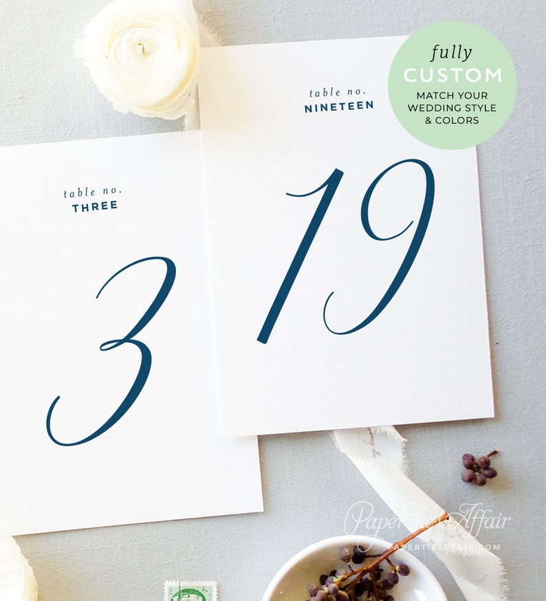 Peut inclure: Deux cartes de num&eacute;ro de table blanches avec une calligraphie bleu marine. La carte de gauche indique "table no. THREE" avec le num&eacute;ro "3". La carte de droite indique "table no. NINETEEN" avec le num&eacute;ro "19".