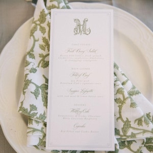 May include: A formal place setting with a white plate, a folded napkin with a green floral pattern, and a menu card. The menu card has a decorative initial and lists a multi-course meal, including a salad, filet of beef, and wedding cake.