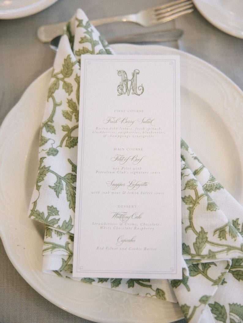 May include: A formal place setting with a white plate, a folded napkin with a green floral pattern, and a menu card. The menu card has a decorative initial and lists a multi-course meal, including a salad, filet of beef, and wedding cake.
