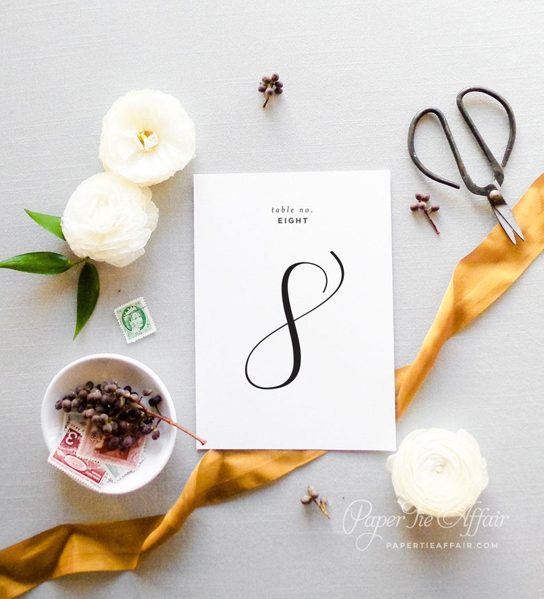 Peut inclure: Carte blanche avec calligraphie noire pour le num&eacute;ro de table huit. La carte est entour&eacute;e de fleurs blanches, d'un ruban dor&eacute; et d'autres &eacute;l&eacute;ments d&eacute;coratifs.