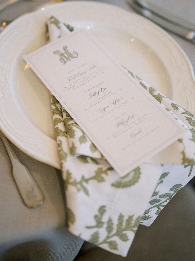 May include: A formal place setting featuring a white plate, a folded napkin with a green floral pattern, and a menu card. The menu card has a decorative initial and a list of courses. A silver fork is visible.