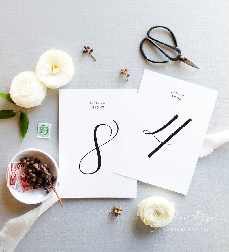 Peut inclure: Deux cartes blanches avec des num&eacute;ros en calligraphie noire, num&eacute;ro de table huit et num&eacute;ro de table quatre, sur un fond gris clair avec des fleurs blanches et une paire de ciseaux.