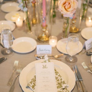 May include: Elegant table setting with a light gray tablecloth, white plates, and silverware. A menu card sits atop a folded napkin with a green floral pattern. Tall glass vases hold pink candles and peach roses, creating a sophisticated ambiance.