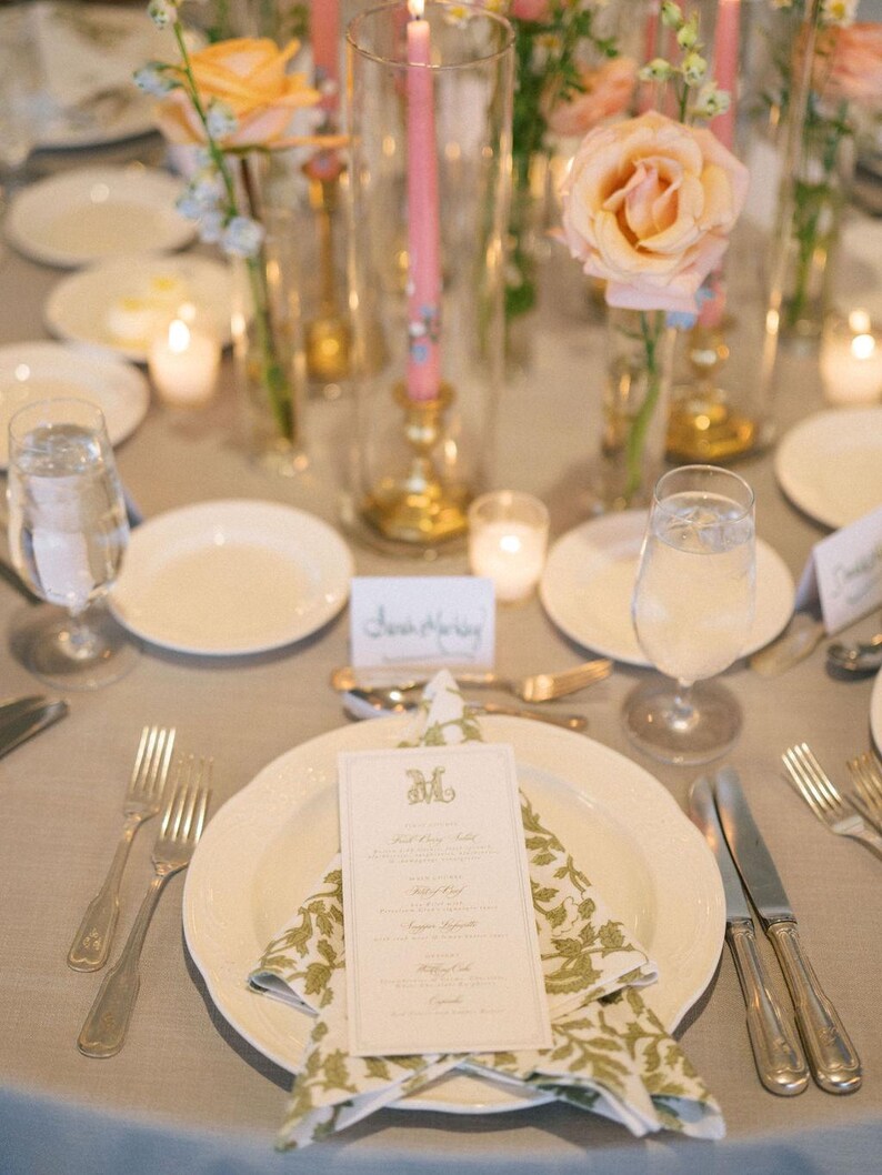 May include: Elegant table setting with a light gray tablecloth, white plates, and silverware. A menu card sits atop a folded napkin with a green floral pattern. Tall glass vases hold pink candles and peach roses, creating a sophisticated ambiance.
