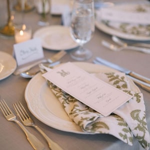 May include: Elegant table setting with a white plate, a folded napkin with a green floral pattern, and a menu card. Silverware and a water glass are also visible. The table is covered with a gray tablecloth.
