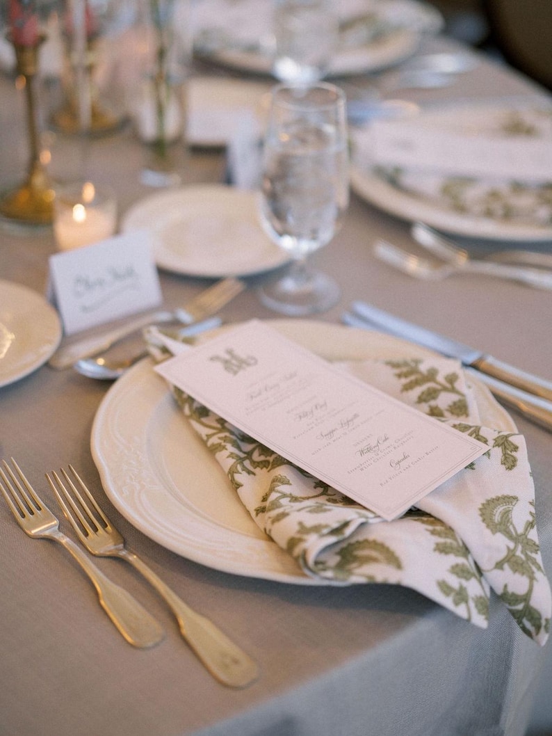 May include: Elegant table setting with a white plate, a folded napkin with a green floral pattern, and a menu card. Silverware and a water glass are also visible. The table is covered with a gray tablecloth.