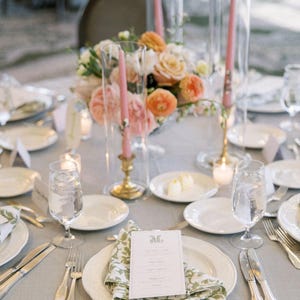 May include: Elegant table setting with a gray tablecloth, white plates, and silverware. A floral centerpiece with peach and white roses sits in the middle. Pink candles in glass holders and printed menu cards complete the look.