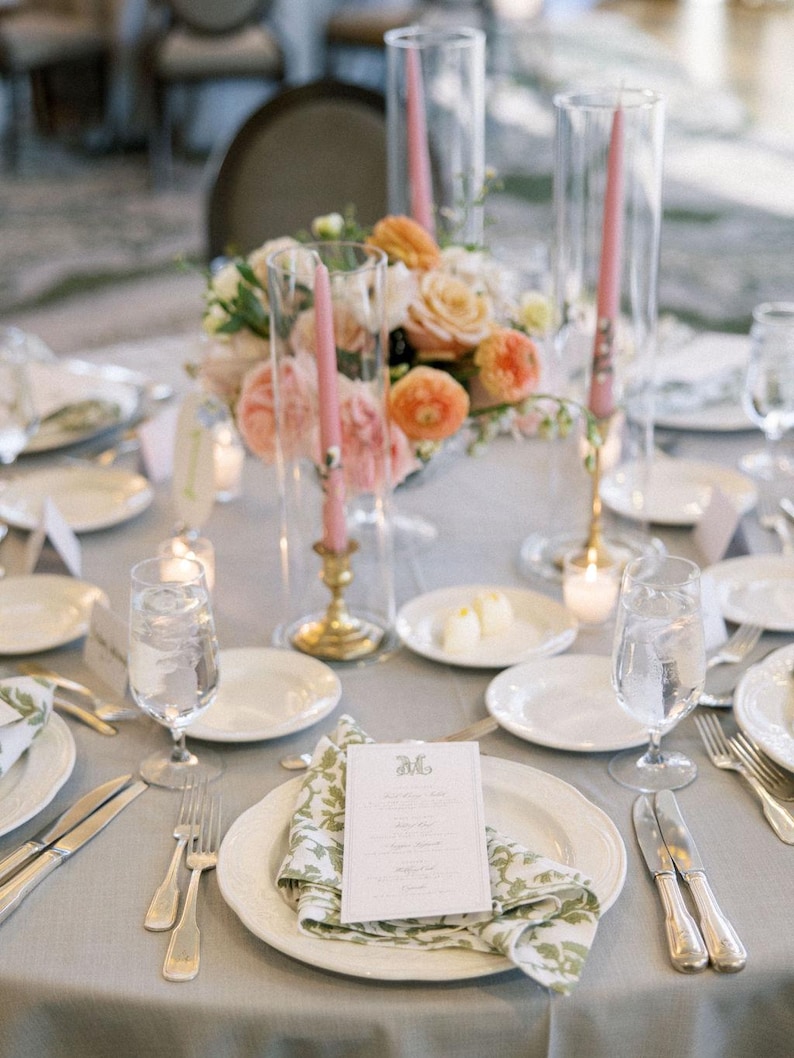 May include: Elegant table setting with a gray tablecloth, white plates, and silverware. A floral centerpiece with peach and white roses sits in the middle. Pink candles in glass holders and printed menu cards complete the look.