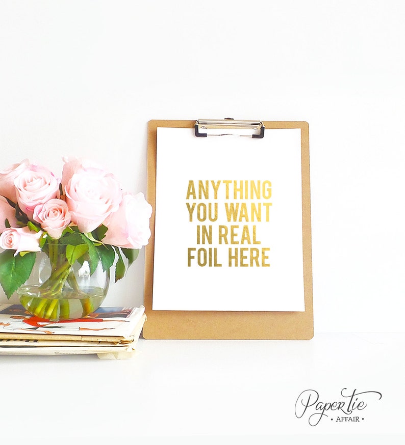 以下が含まれることがあります： 「ANYTHING YOU WANT IN REAL FOIL HERE」と書かれた金色の箔文字が入った、茶色のクリップボードに入った白い紙。ピンクのバラが入ったガラスの花瓶と雑誌の束が左側にあります。