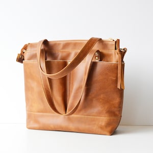 Puede incluir: Un bolso tote grande de cuero marr&oacute;n con dos asas y m&uacute;ltiples bolsillos. El bolso tiene forma rectangular con fondo plano y cierre de cremallera. El cuero tiene un aspecto envejecido y el bolso parece nuevo.