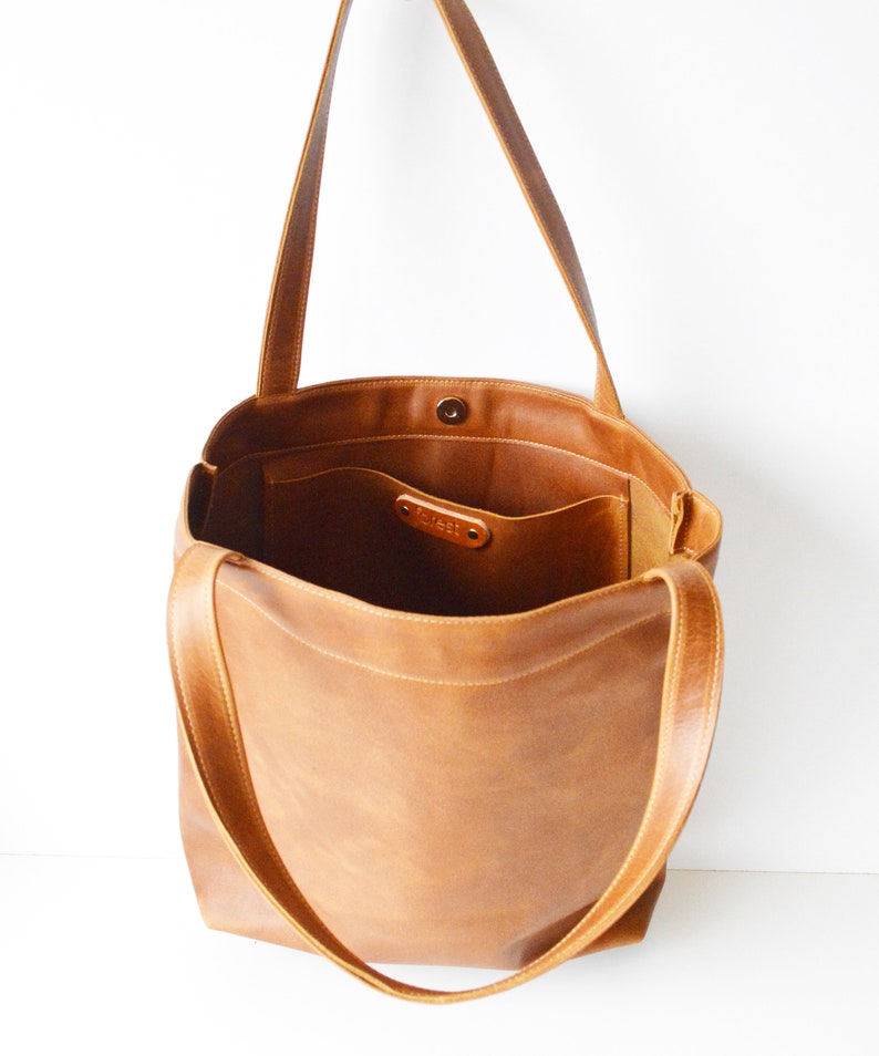 May include: A light brown leather tote bag with long shoulder straps. The bag has an open top with a magnetic snap closure and an interior pocket. The word "forest" is embossed on a small leather tag inside the bag.