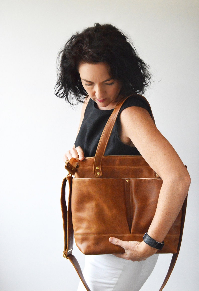 Puede incluir: Un bolso tote de cuero marr&oacute;n con una correa para el hombro. El bolso tiene forma rectangular con un bolsillo frontal y remaches dorados. El bolso lo sostiene una persona que lleva una camiseta negra y pantalones blancos.