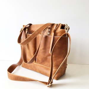 Puede incluir: Un bolso tote de cuero marr&oacute;n con dos bolsillos delanteros y una correa larga ajustable para el hombro. El bolso tiene dos asas superiores y herrajes dorados. El cuero tiene un aspecto envejecido.