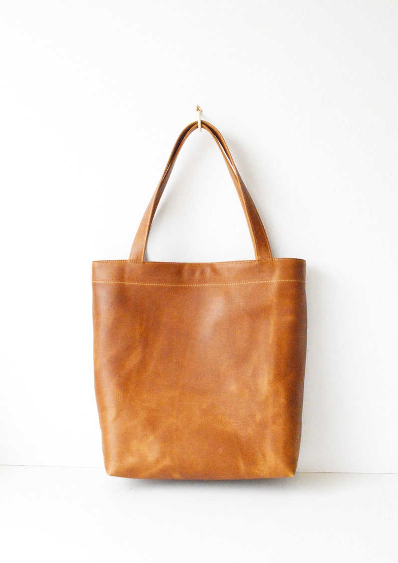 May include: A brown leather tote bag with two long handles, hanging against a white background. The bag has a rectangular shape with a simple design and visible stitching along the top.