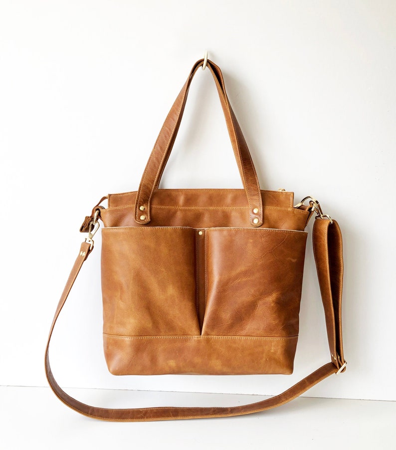 Puede incluir: Un bolso tote de cuero marr&oacute;n con dos bolsillos delanteros y una correa larga y ajustable para el hombro. El bolso tiene dos asas superiores y herrajes dorados. El bolso cuelga sobre un fondo blanco.