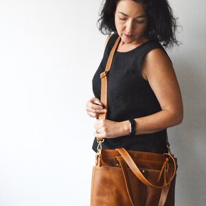 Puede incluir: Un bolso de hombro de cuero marr&oacute;n con una correa larga ajustable. El bolso tiene forma rectangular con m&uacute;ltiples bolsillos y herrajes dorados. El bolso se lleva al hombro.