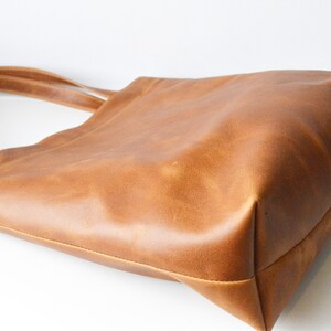 May include: A brown leather tote bag with two shoulder straps. The bag is made of a smooth, textured leather and has a rectangular shape with a flat bottom. The bag is empty and sits on a white surface.