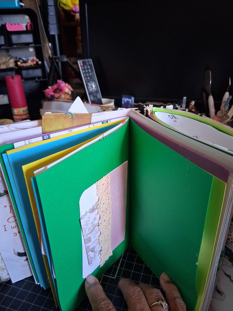 Pu&ograve; includere: Un album di ritagli fatto a mano con pagine aperte che presentano inserti in cartoncino verde, giallo e blu vivaci. Le pagine includono un piccolo cartoncino bianco con un bordo in pizzo beige e un'illustrazione stampata.