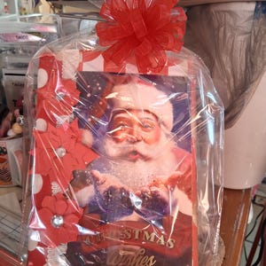 May include: A Christmas gift set wrapped in clear cellophane and topped with a large red bow. The gift includes a Christmas card featuring Santa Claus and the words "Christmas Wishes". The card is decorated with red and white polka dots and red paper flowers.