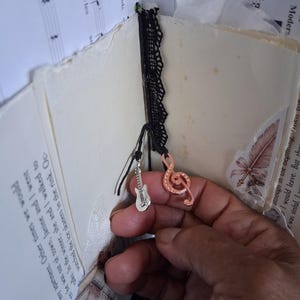 Pu&ograve; includere: Un diario fatto a mano con pagine color crema contenenti testo stampato e notazione musicale. Un nastro di pizzo nero funge da segnalibro, decorato con due ciondoli in metallo: una chitarra elettrica argentata e una chiave di violino ramata.