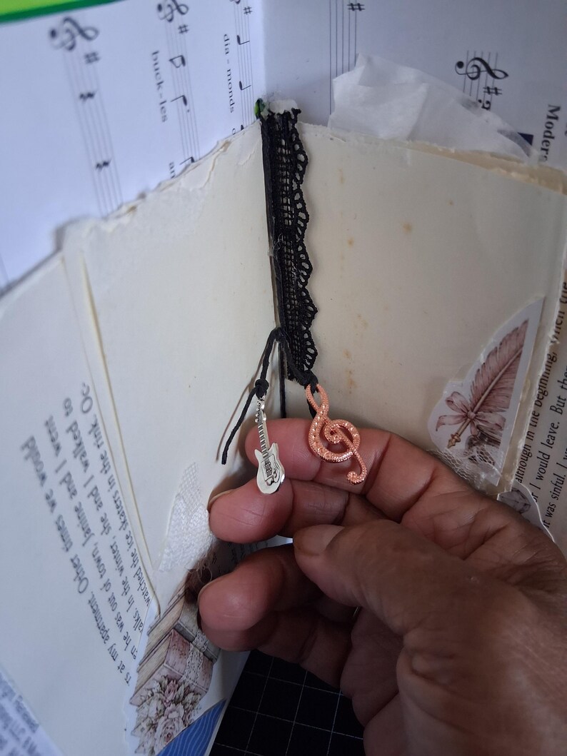 Pu&ograve; includere: Un diario fatto a mano con pagine color crema contenenti testo stampato e notazione musicale. Un nastro di pizzo nero funge da segnalibro, decorato con due ciondoli in metallo: una chitarra elettrica argentata e una chiave di violino ramata.