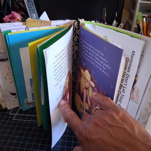 Pu&ograve; includere: Un album di ritagli fatto a mano con pagine multicolori, tra cui carta giallo brillante e blu. Una pagina mostra un'illustrazione stampata di una persona bionda con un cappello da cowboy e una chitarra, con il testo Mammas Don't Let Your Babies Grow Up to Be Cowboys.