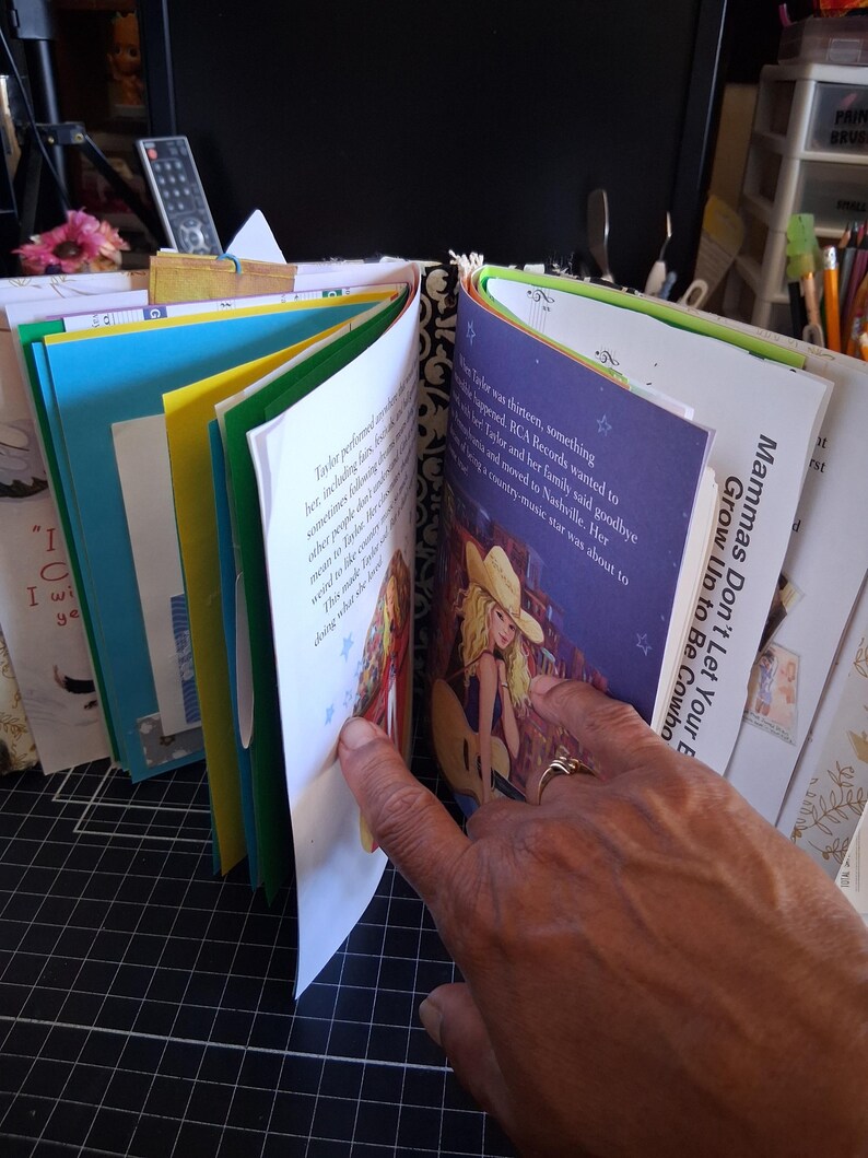 Pu&ograve; includere: Un album di ritagli fatto a mano con pagine multicolori, tra cui carta giallo brillante e blu. Una pagina mostra un'illustrazione stampata di una persona bionda con un cappello da cowboy e una chitarra, con il testo Mammas Don't Let Your Babies Grow Up to Be Cowboys.