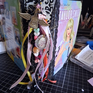 Pu&ograve; includere: Un libro biografico Little Golden Book su Taylor Swift riadattato, con dorso in damasco bianco e nero decorato con pizzo nero, un ciondolo a forma di chitarra alata color oro, un pendente circolare glitterato e vari nastri e perline color pastello.