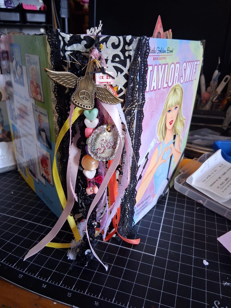 Pu&ograve; includere: Un libro biografico Little Golden Book su Taylor Swift riadattato, con dorso in damasco bianco e nero decorato con pizzo nero, un ciondolo a forma di chitarra alata color oro, un pendente circolare glitterato e vari nastri e perline color pastello.