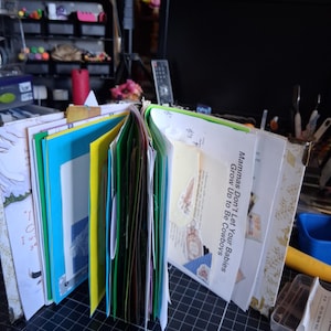Pu&ograve; includere: Un junk journal fatto a mano con pagine colorate, inclusi inserti in carta blu brillante, verde e giallo. Il libro aperto mostra vari ritagli di carta, efimeri e una pagina con il testo stampato, "Mammas Don't Let Your Babies Grow Up to Be Cowboys."
