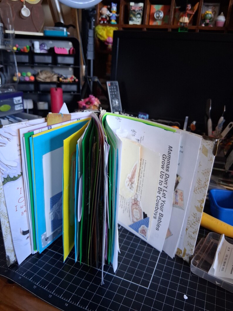 Pu&ograve; includere: Un junk journal fatto a mano con pagine colorate, inclusi inserti in carta blu brillante, verde e giallo. Il libro aperto mostra vari ritagli di carta, efimeri e una pagina con il testo stampato, "Mammas Don't Let Your Babies Grow Up to Be Cowboys."