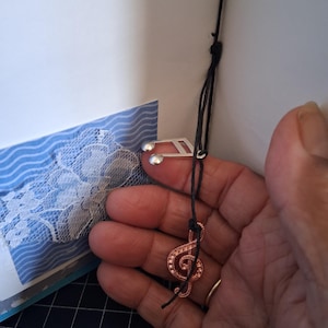 Pu&ograve; includere: Un ciondolo a forma di nota musicale color argento e un ciondolo a forma di chiave di violino color rame attaccati a sottili cordini neri. I ciondoli sono sospesi vicino a un inserto di carta blu e bianca a onde con pizzo bianco.