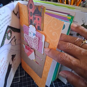 Pu&ograve; includere: Una pagina di scrapbooking con uno sfondo arancione a motivi e una tasca in cartoncino marrone. La tasca contiene una farfalla rosa fustellata, un piccolo ritaglio a forma di casa e un'etichetta decorativa in un libro illustrato.