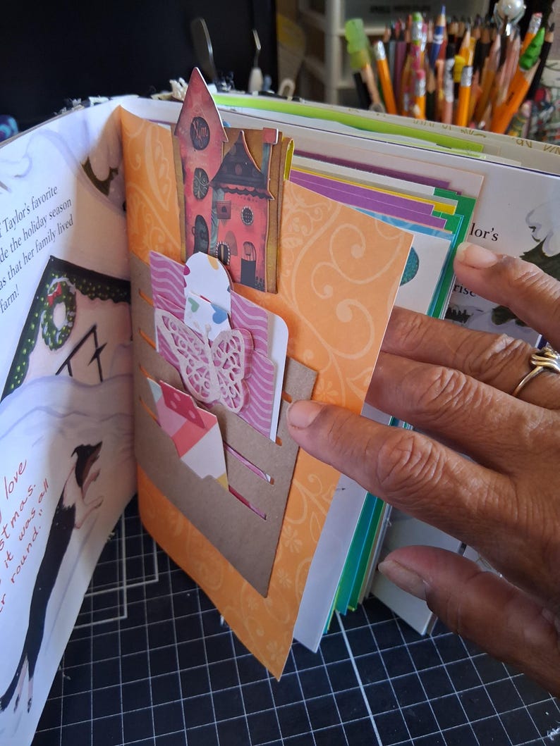 Pu&ograve; includere: Una pagina di scrapbooking con uno sfondo arancione a motivi e una tasca in cartoncino marrone. La tasca contiene una farfalla rosa fustellata, un piccolo ritaglio a forma di casa e un'etichetta decorativa in un libro illustrato.