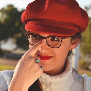 May include: A woman wearing a red newsboy cap, brown tortoiseshell glasses, and a white turtleneck sweater with a brown and white pattern. She is wearing a silver ring with turquoise stones on her finger.