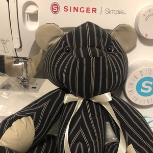 May include: A black and white striped teddy bear with a white satin ribbon around its neck. The bear is sitting on a white surface.