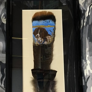 May include: A framed painting of a brown and white dog on a feather. The dog is painted in a realistic style and is looking to the left. The feather is brown and white and has a blue and green background. The painting is signed "R.D.S."