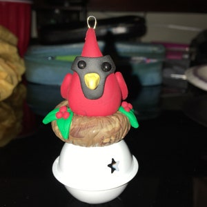 May include: A red cardinal bird ornament with black eyes and a yellow beak, sitting in a brown nest with green leaves and red berries, on top of a white bell with a star cutout.