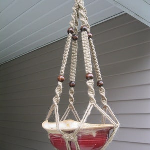 May include: A handmade macrame plant hanger with a red ceramic pot. The beige rope is knotted and features dark brown wooden beads. The pot has a cream-colored rim and is suspended from a metal ring.