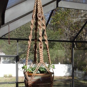 May include: A handcrafted macrame plant hanger made of natural jute rope, featuring wooden beads and a tassel. The hanging planter holds a ceramic pot with green plants, perfect for indoor or outdoor decor.