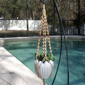 May include: A macrame plant hanger with a white ceramic pot filled with green plants. The beige macrame has wooden beads and a tassel. The plant holder is suspended from a black metal stand near a pool.