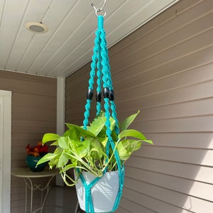 May include: A turquoise macrame plant hanger with a white pot holding a green leafy plant. The hanger has black beads and a tassel. The plant is suspended from a metal hook.