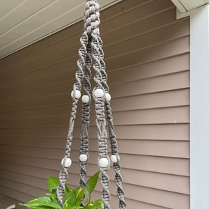 May include: A gray macrame plant hanger with white beads, holding a potted plant with green and yellow leaves. The plant hanger is suspended from a white ceiling, creating a decorative and functional display for indoor or outdoor use.