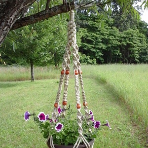 May include: A macrame plant hanger with a brown pot filled with purple and white petunias. The beige rope hanger is adorned with wooden beads and hangs from a tree branch in a grassy outdoor setting.