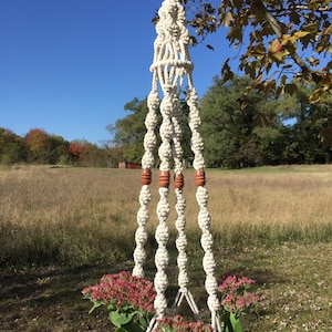 May include: A cream-colored macrame plant hanger with a pot of pink flowers. The hanger is suspended from a tree branch against a backdrop of a field and blue sky. The macrame design includes wooden beads.