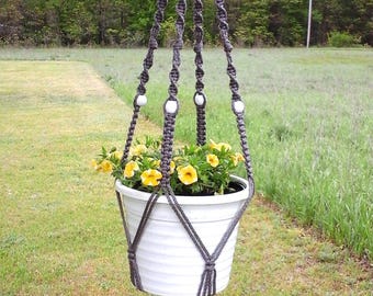 May include: A gray macrame plant hanger with a white pot filled with yellow flowers. The hanger is suspended from a tree branch and features white beads. The pot is hanging in a grassy outdoor setting.