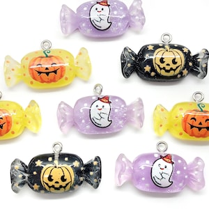 May include: A collection of Halloween-themed candy-shaped charms. The charms feature a smiling jack-o'-lantern, a friendly ghost, and a wrapped candy design. The candies are in yellow, purple, and black with silver-colored loops for attaching to jewelry or other crafts.