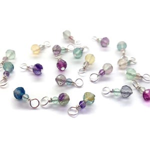 May include: A collection of 18 small, colorful gemstone beads with silver wire loops. The beads are various shades of green, blue, purple, and yellow.
